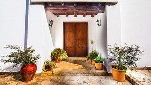 Um edifício branco com porta de madeira, plantas em vasos e um caminho de pedra que leva à entrada.