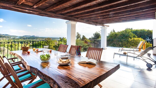 Uma mesa de jantar de madeira com cadeiras, vista para o campo e uma varanda com guarda-corpo.