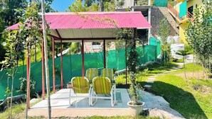 Een overdekt terras met stoelen en een tafel, omgeven door groen en een gebouw op de achtergrond.