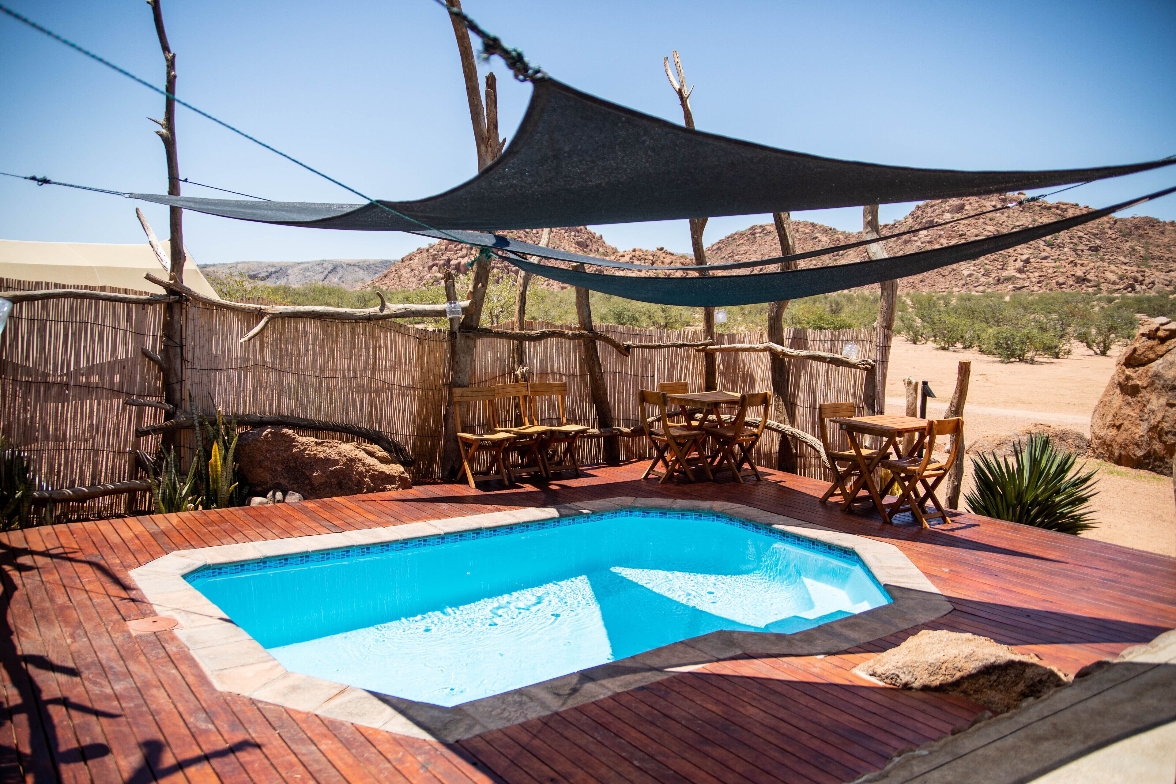 Una terraza de madera con piscina, rodeada de mobiliario de madera y una zona con sombra.