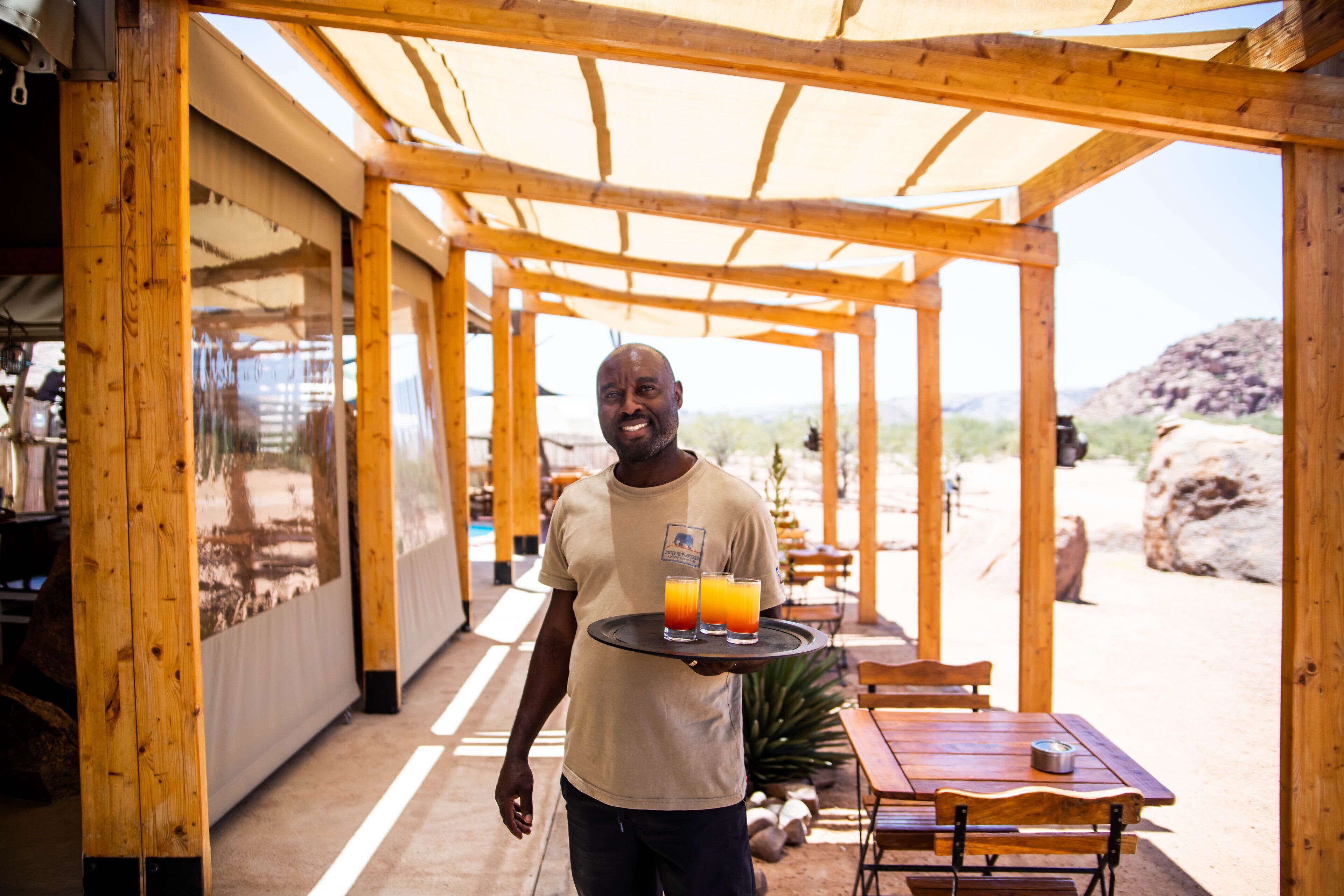 A wooden structure with tables and chairs, a person carrying a tray with drinks, and a desert landscape in the background.