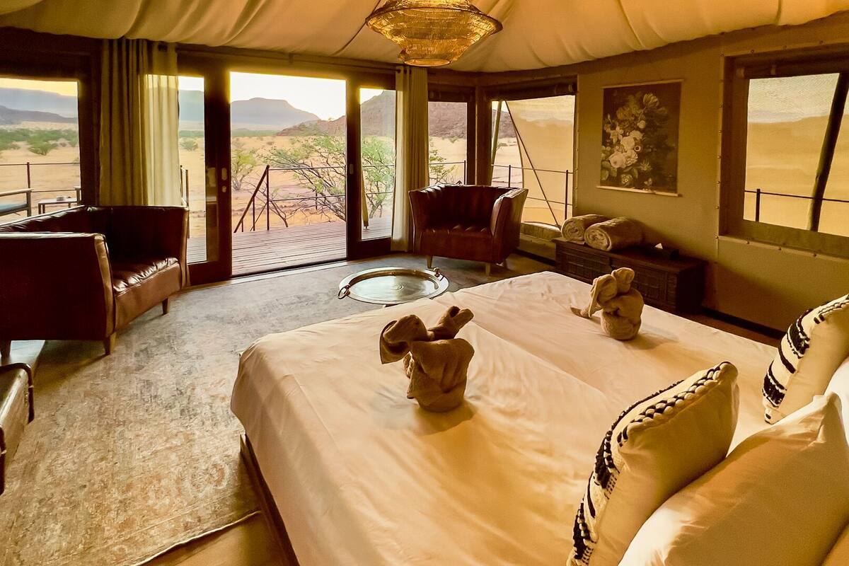 A room with a bed, two chairs, a dining table, and a view of the desert.