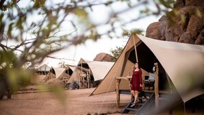 Campaamento en el desierto con varias tiendas de campaña, una mujer con vestido rojo parada en una escalera de madera y un paisaje rocoso al fondo.
