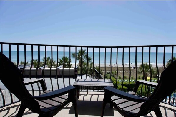 A balcony with two chairs and a small table, overlooking a beach with palm trees and a clear sky.