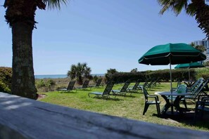 Un espace de détente aménagé au bord de la plage, avec plusieurs rangées de chaises longues et de tables, dont certaines sont à l’ombre de parasols, et qui offre une vue sur l’océan.