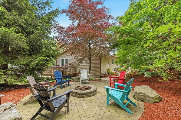 A patio area with Adirondack chairs, a fire pit, and a house in the background.