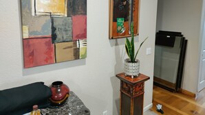 A living room with a modern abstract painting, a small wooden table with a vase and a bottle, and a potted plant.