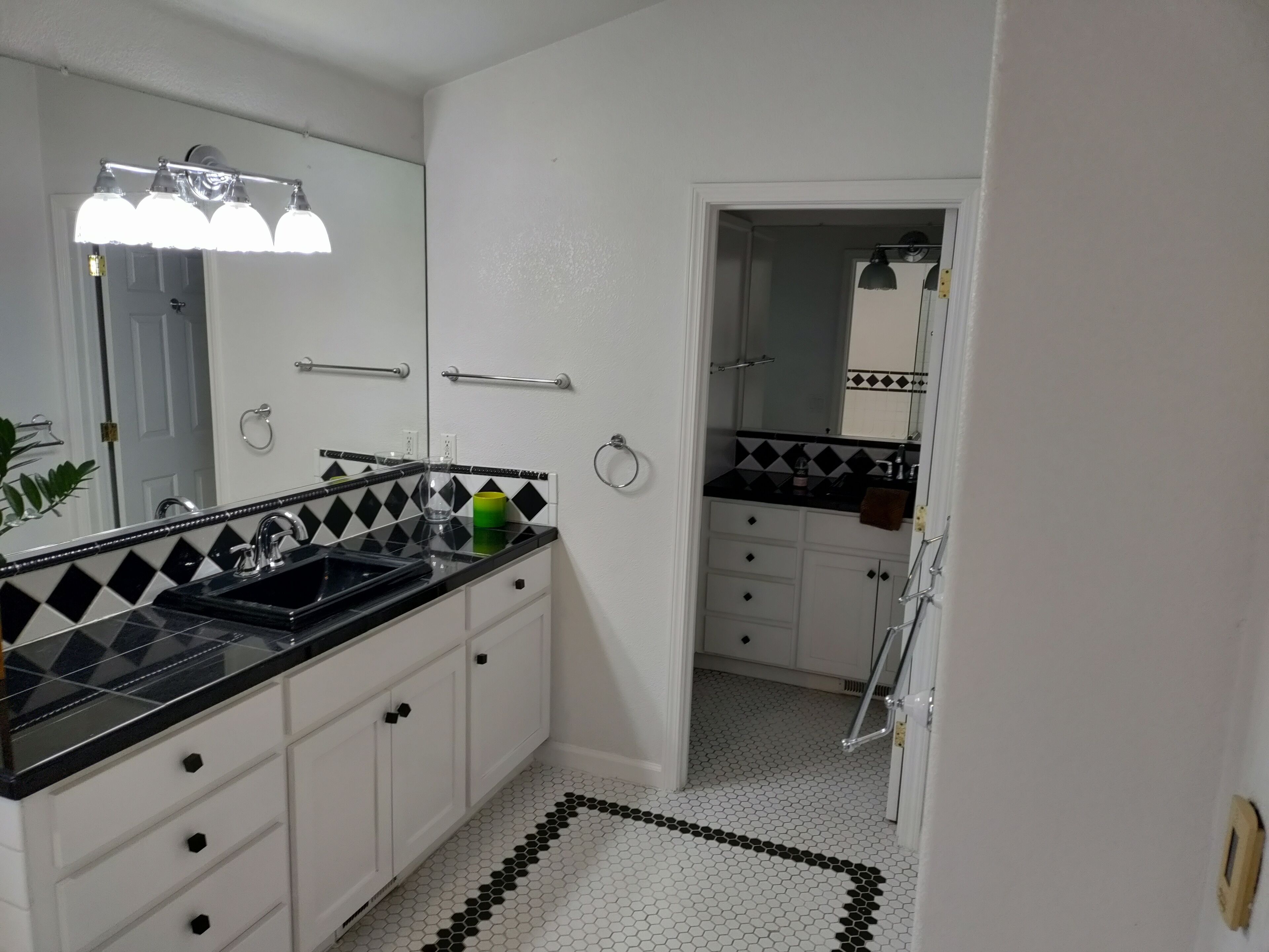 Un baño con suelo de baldosas blanco y negro, un lavabo negro, armarios blancos y un espejo con una lámpara arriba.