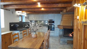 A wooden dining table with chairs, a vase with dried branches, a kitchen with modern appliances, and a stone fireplace.
