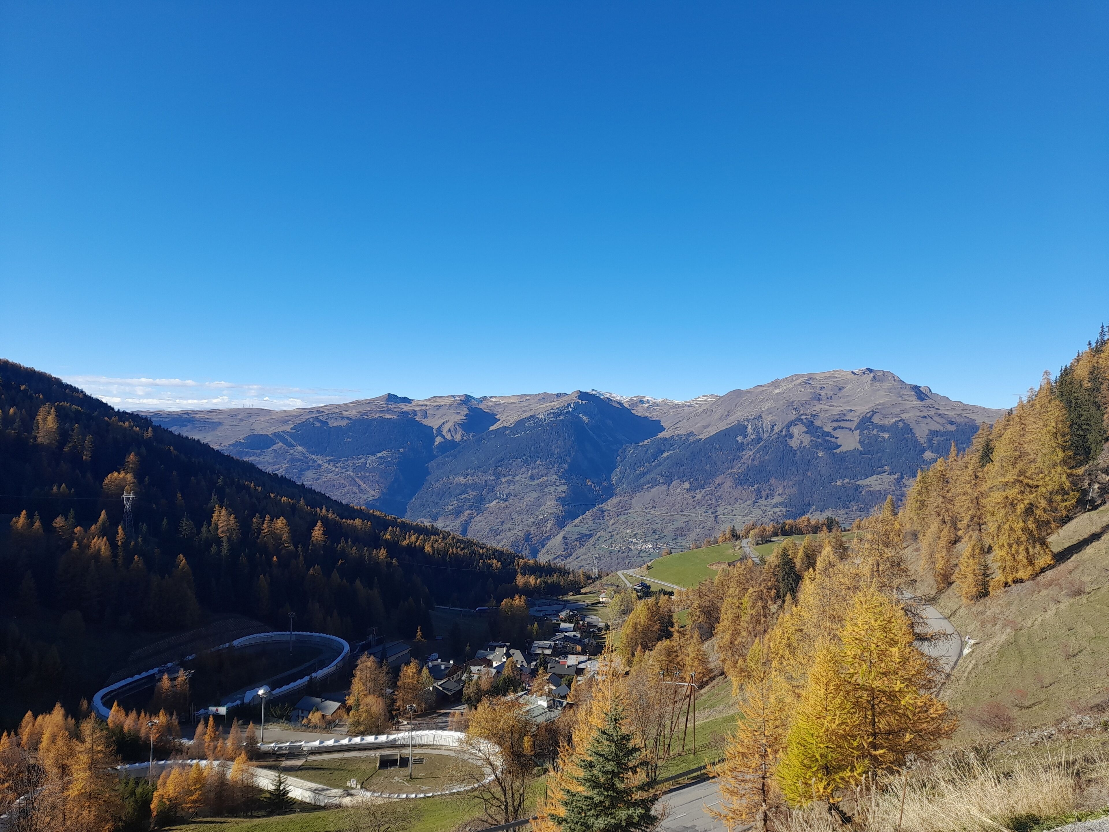 Eine gebirgige Landschaft mit einer kurvenreichen Straße, einer kleinen Siedlung und blauem Himmel.