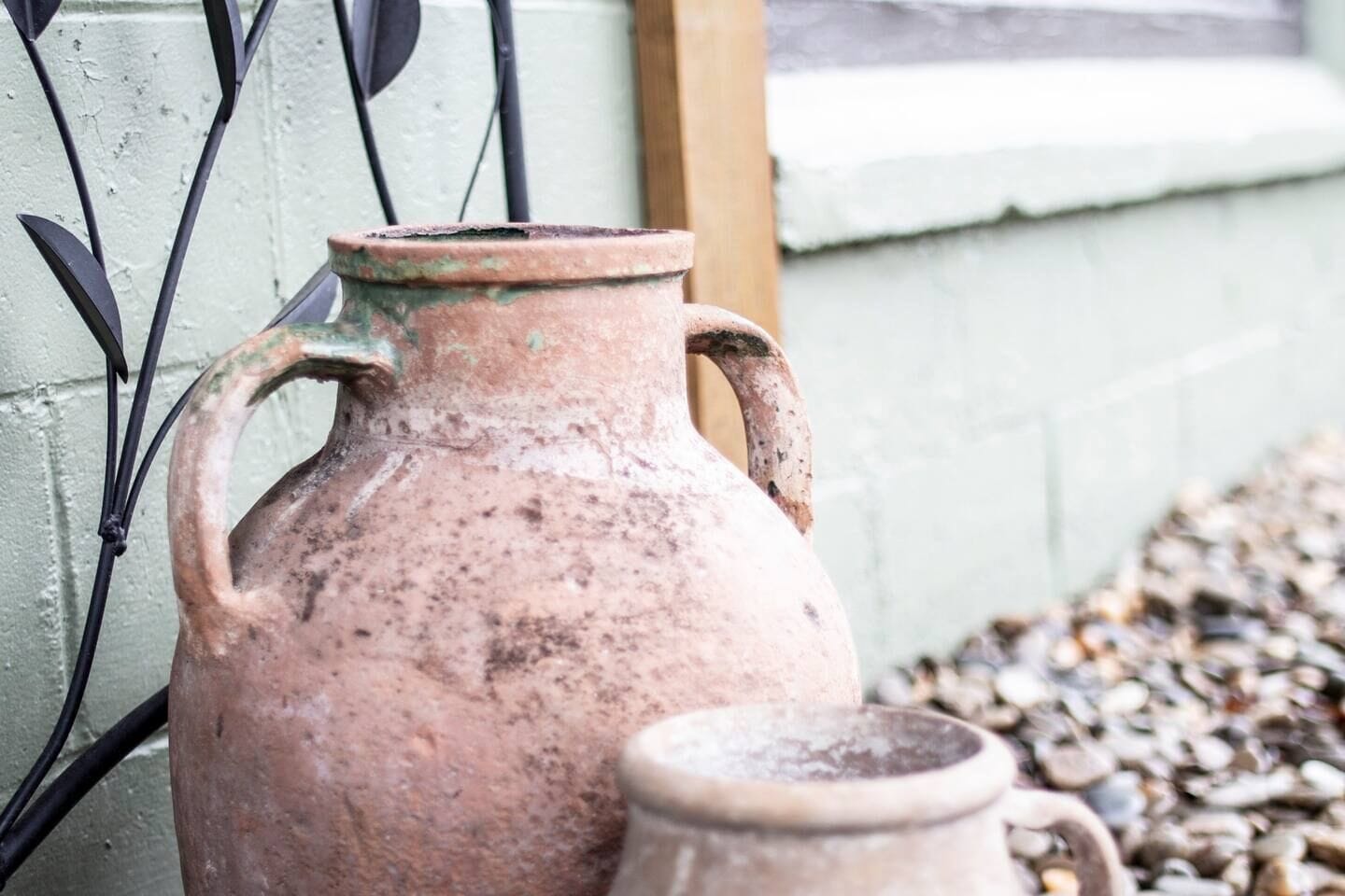 A large, rustic clay pot with handles, placed against a wall with a window and a metal fence.