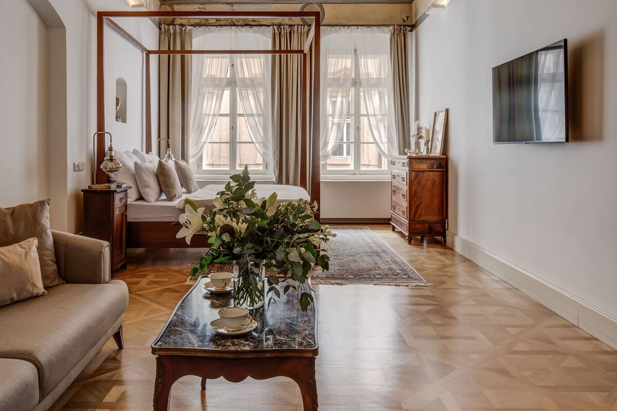 A spacious room with a wooden floor, a four-poster bed, a sitting area with a sofa, a coffee table, and a television mounted on the wall.