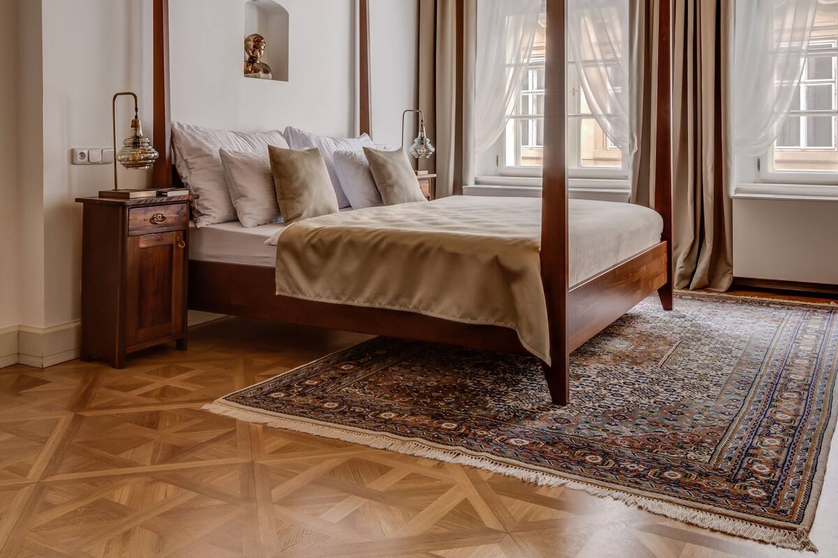 A four-poster bed with a wooden frame, a nightstand with a lamp, and a patterned rug on a parquet floor.