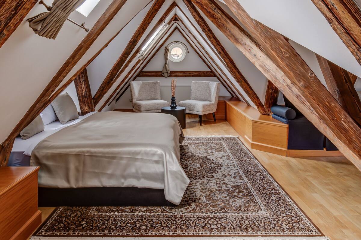A bedroom with a sloped ceiling, wooden beams, a bed, a chair, a small table, and a rug.