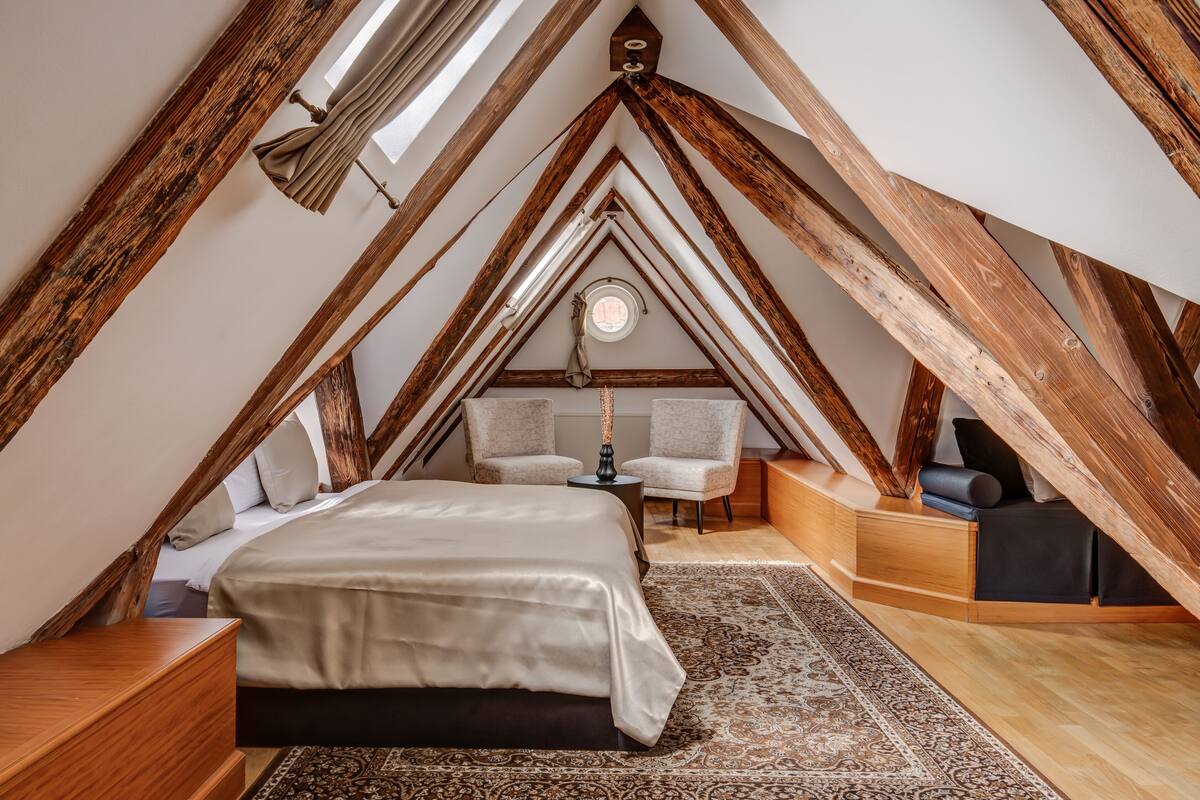 A bedroom with a sloped ceiling, wooden beams, a bed, two chairs, a small table, and a rug.