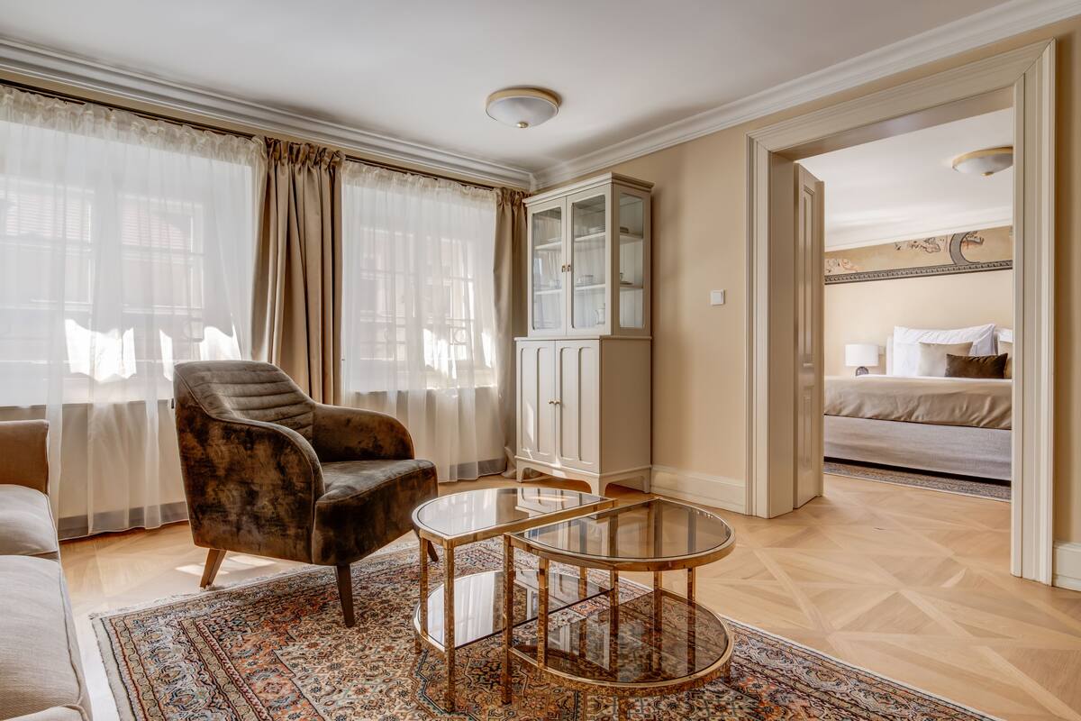 A hotel room with a large window, a plush armchair, a glass-top coffee table, a cabinet, and a bed.