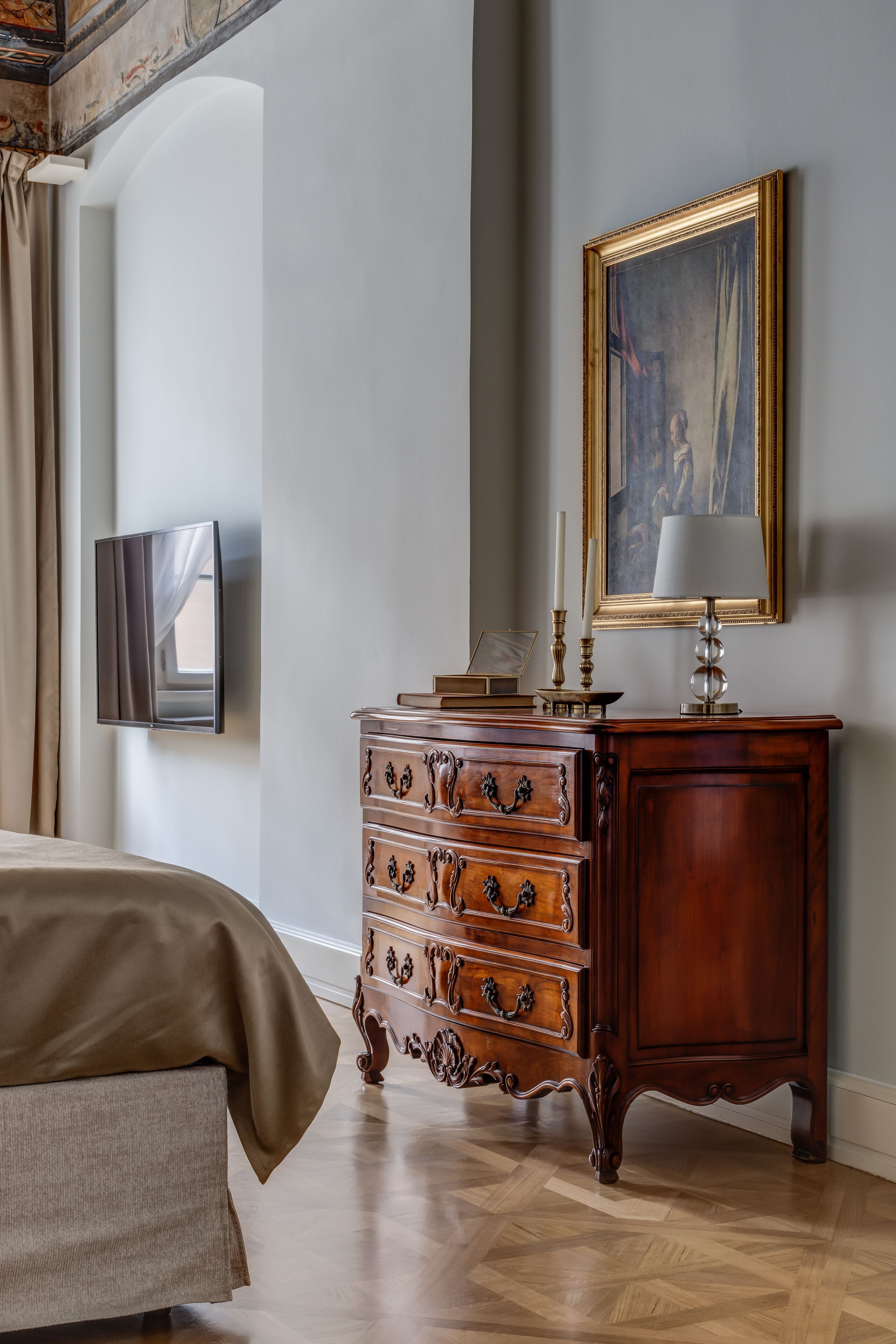 A bedroom with a wooden dresser, a painting on the wall, a television, and a bed with a beige cover.