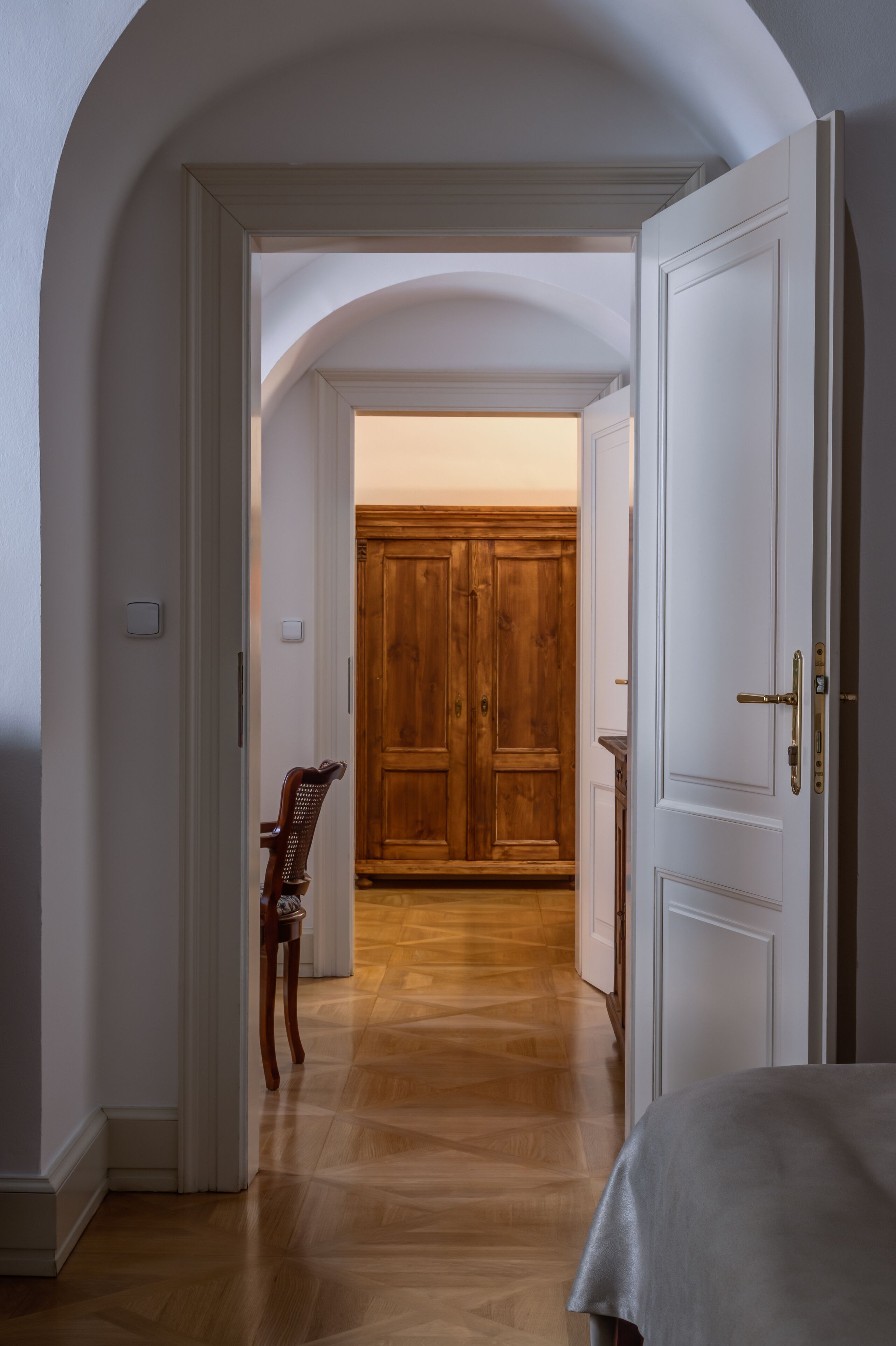 Un pasillo con un armario de madera, una silla y una cama con cabecero blanco.