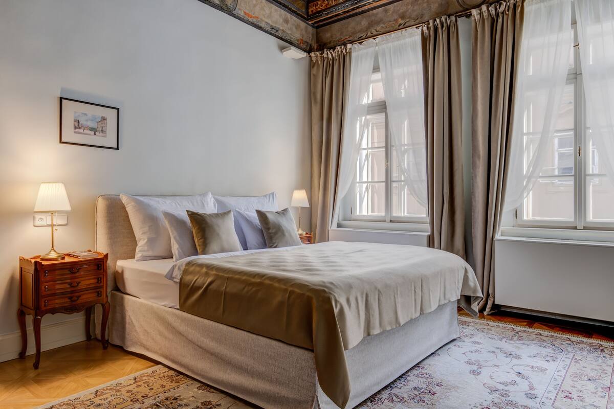 A bedroom with a bed, two bedside tables, a nightstand, a window with curtains, and a framed picture on the wall.