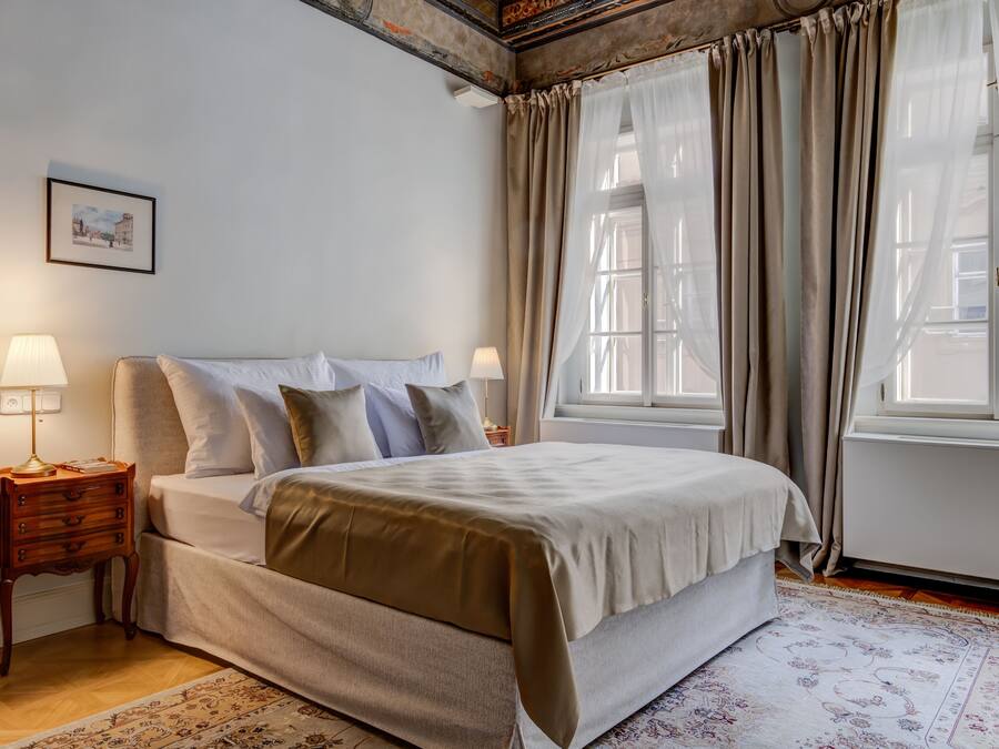 Un dormitorio con cama, dos mesitas de noche, una mesita auxiliar, una ventana con cortinas y un cuadro enmarcado en la pared.