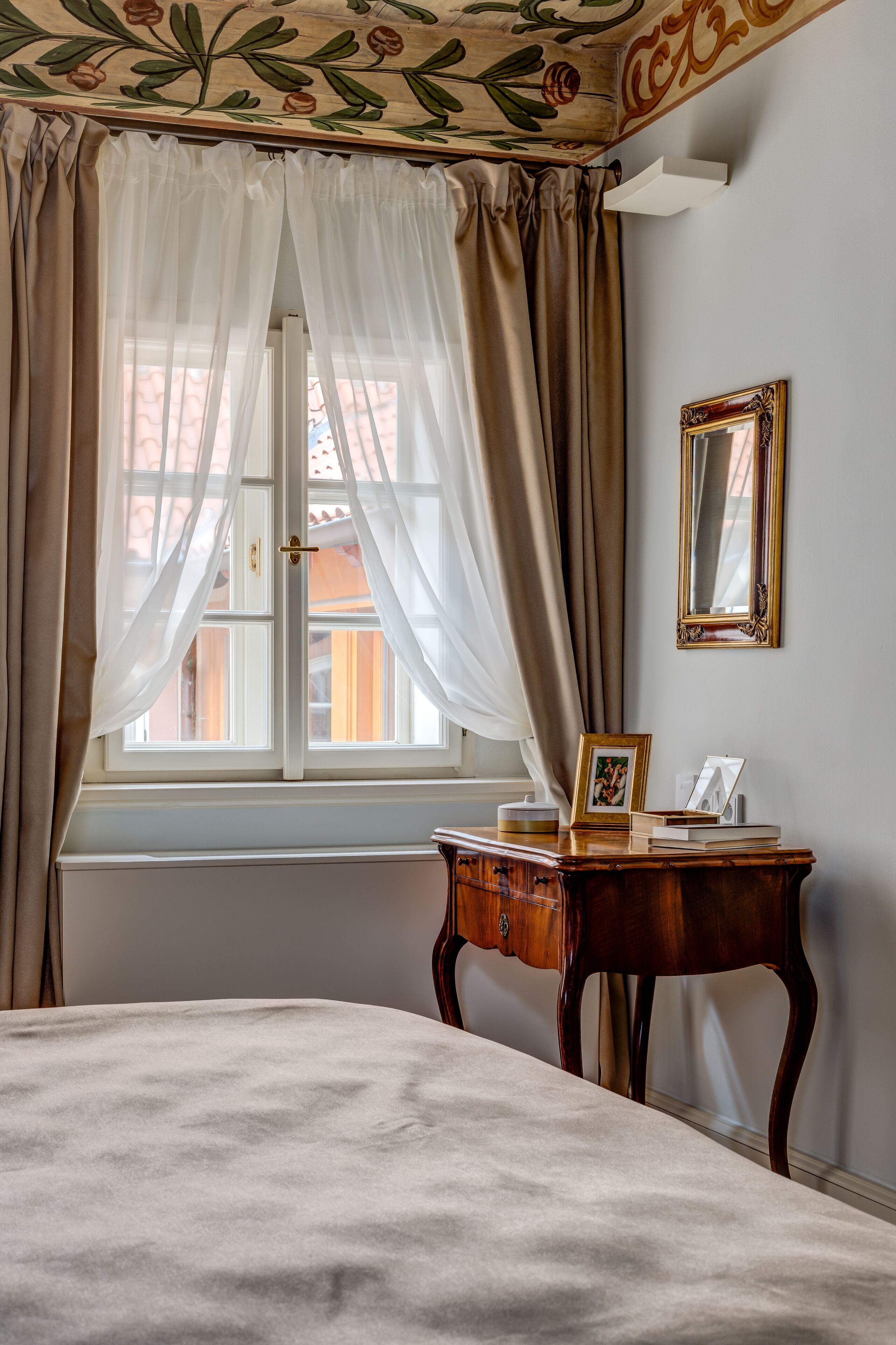 Una habitación con una cama, una mesita de noche de madera, una ventana con cortinas translúcidas y un espejo adosado a la pared.