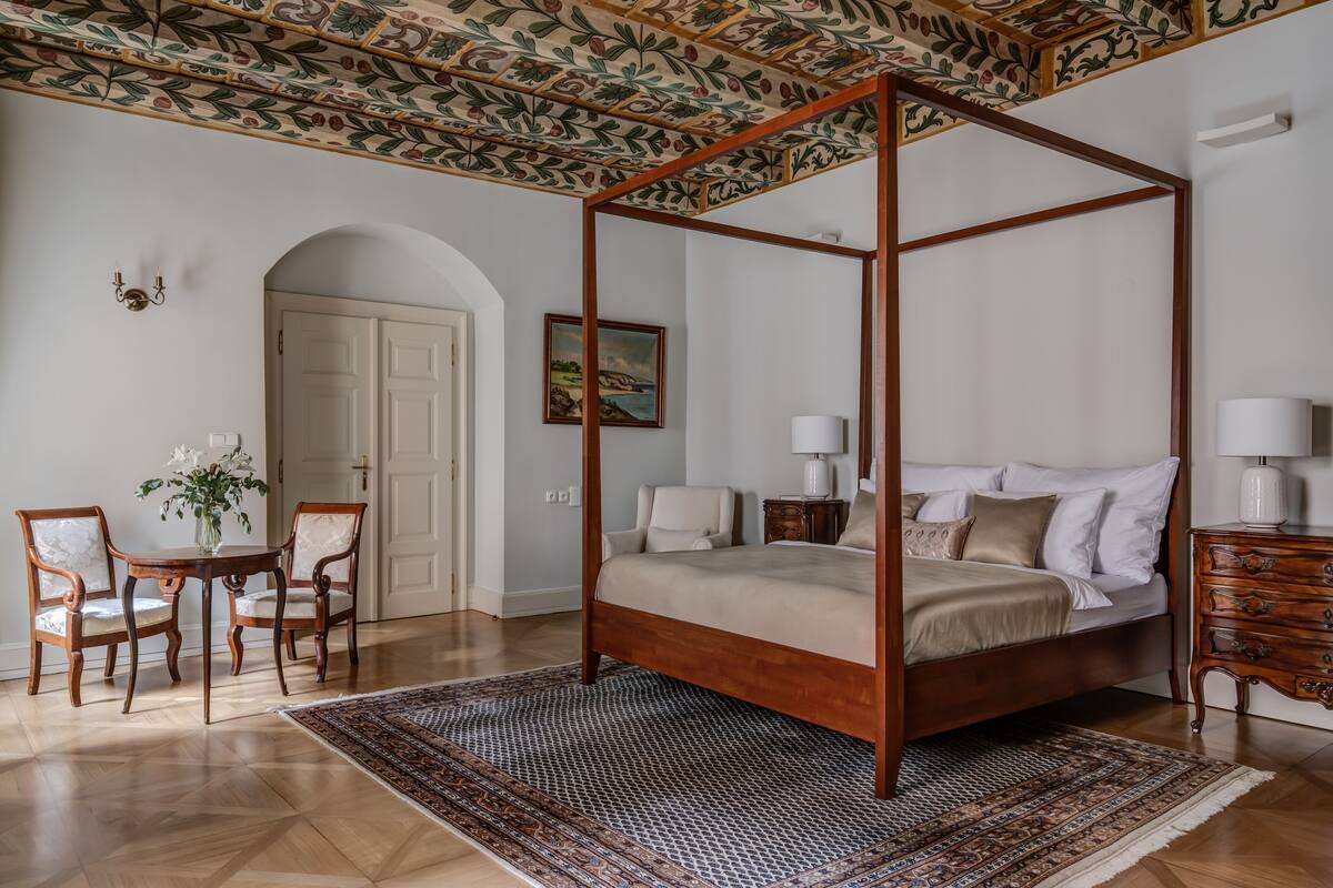 A four-poster bed with a wooden frame, a dining table with chairs, and a patterned rug in a room with a wooden floor and a ceiling with a floral design.