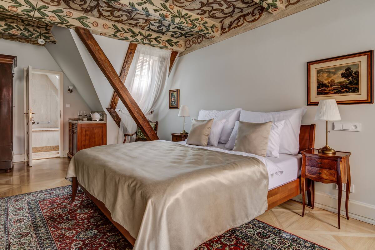 A bedroom with a large bed, wooden beams, a patterned ceiling, and a painting on the wall.