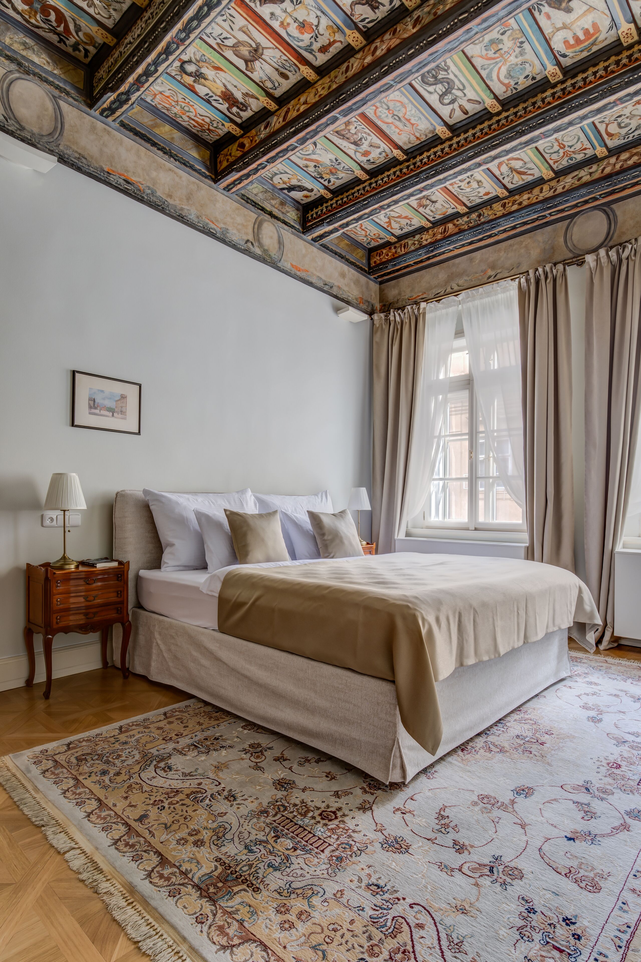 A bedroom with a large bed, a nightstand, a lamp, a window with curtains, and a detailed ceiling.