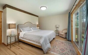 Ein Schlafzimmer mit einem großen Bett, Nachttischlampen, einem kleinen Tisch und Blick ins Freie durch eine Schiebetür.
