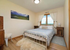 A bedroom with a bed, a chair, a painting, a window with curtains, and a wooden floor.
