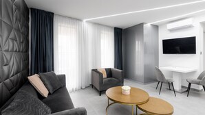 A modern hotel room with a sofa, armchairs, a round table, and a TV mounted on the wall. - Avand Apartments Debrecen (Debrecen)