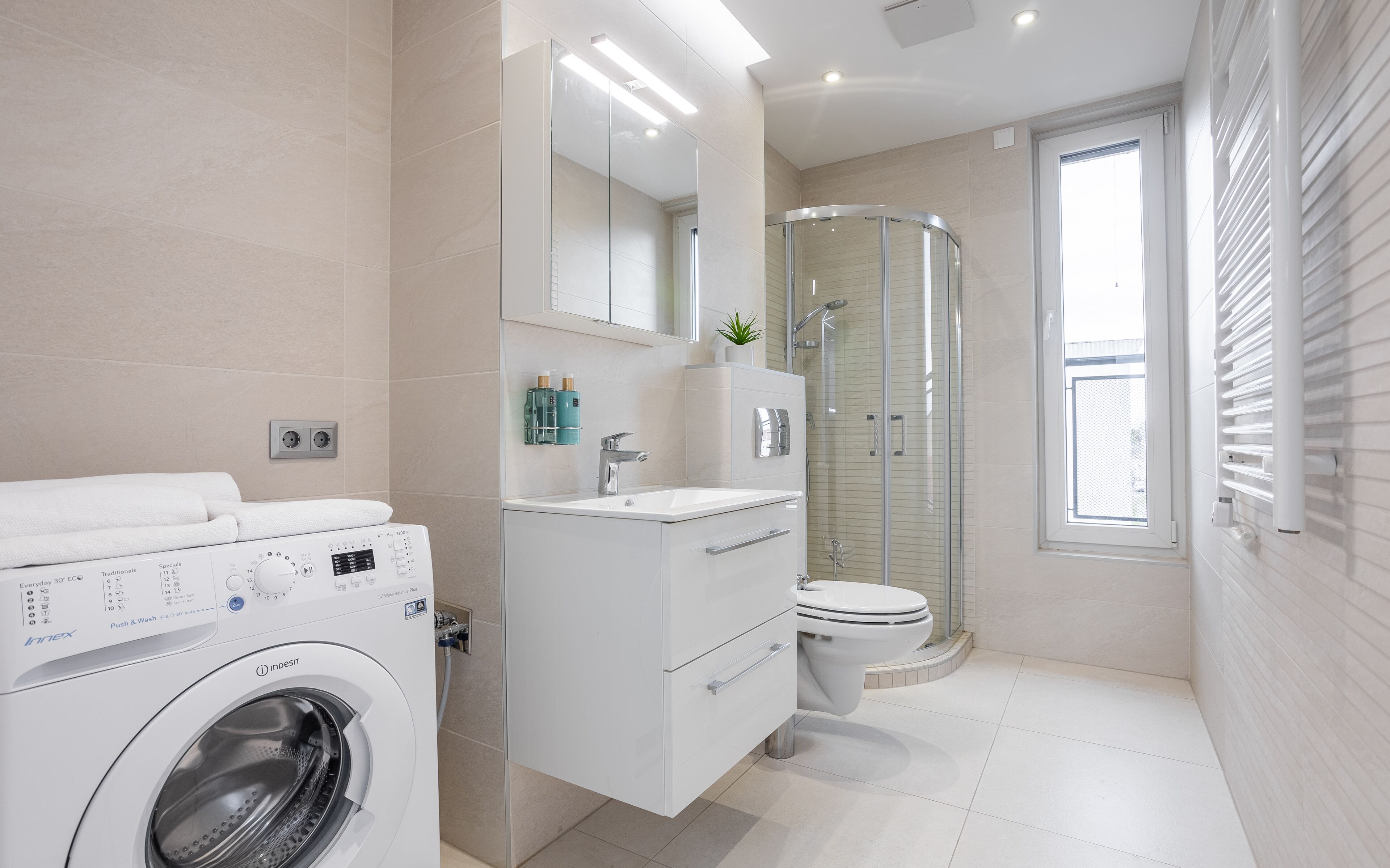 Un bagno moderno con lavatrice, box doccia in vetro, WC e finestra che permette l'illuminazione naturale.