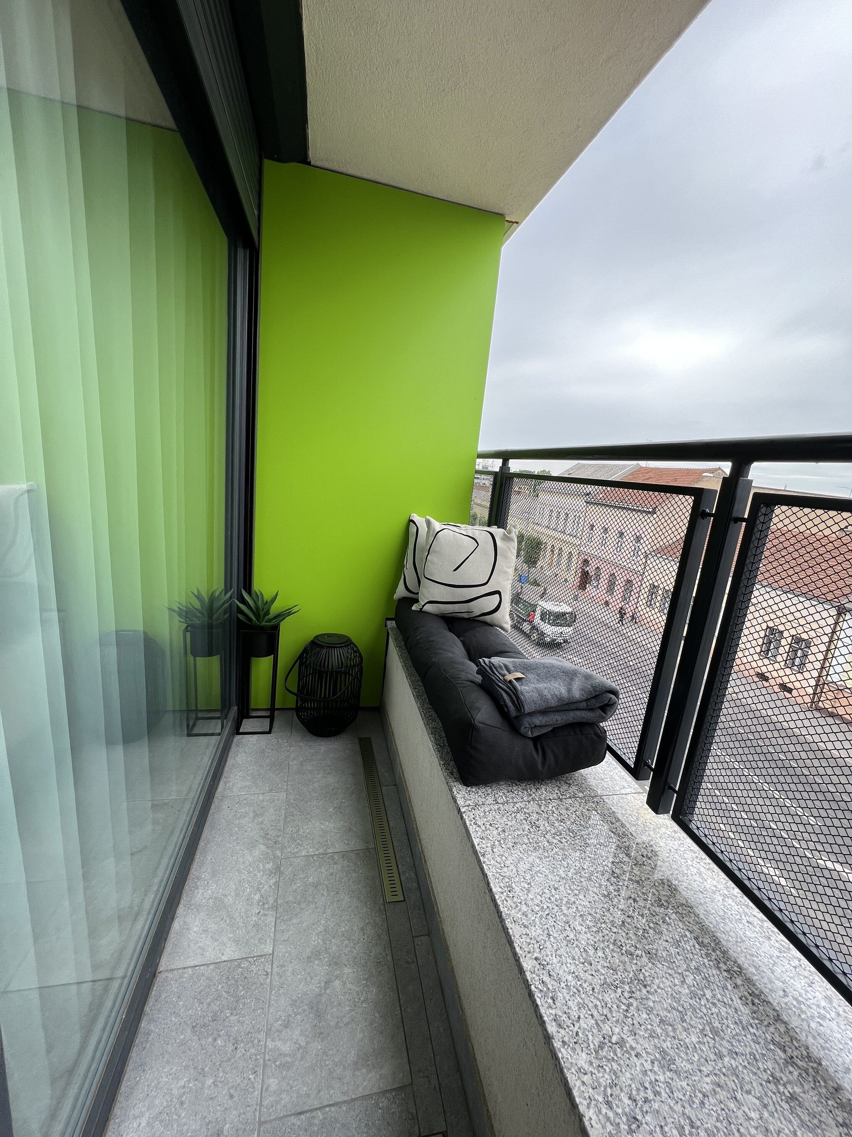 Un balcón con un muro verde, un sofá negro con cojines y una ventana que ofrece vistas a edificios.