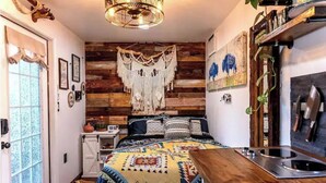 A cozy bedroom with a wooden wall, a bed with patterned bedding, a hanging macrame, and a rustic kitchen area. - Cozy AF Tiny-House Oasis (Tampa)