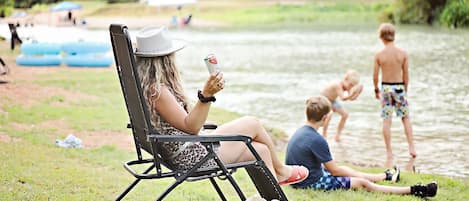 A person relaxing in a chair by a lake, with a bag and a drink on the ground.