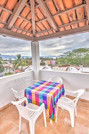 A covered balcony with a table and chairs, a colorful tablecloth, and a view of a residential area with palm trees and buildings.