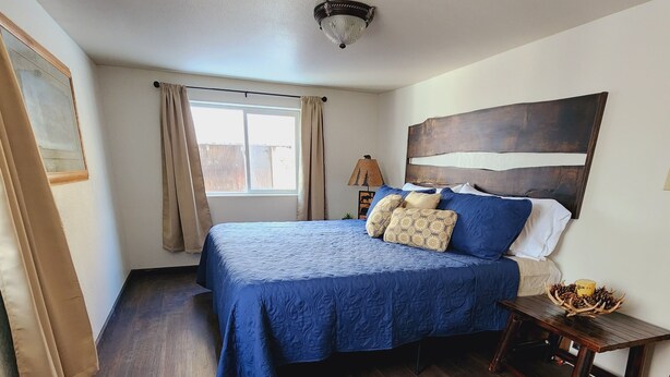 A bedroom with a large bed, wooden flooring, a ceiling light fixture, and a window with curtains.