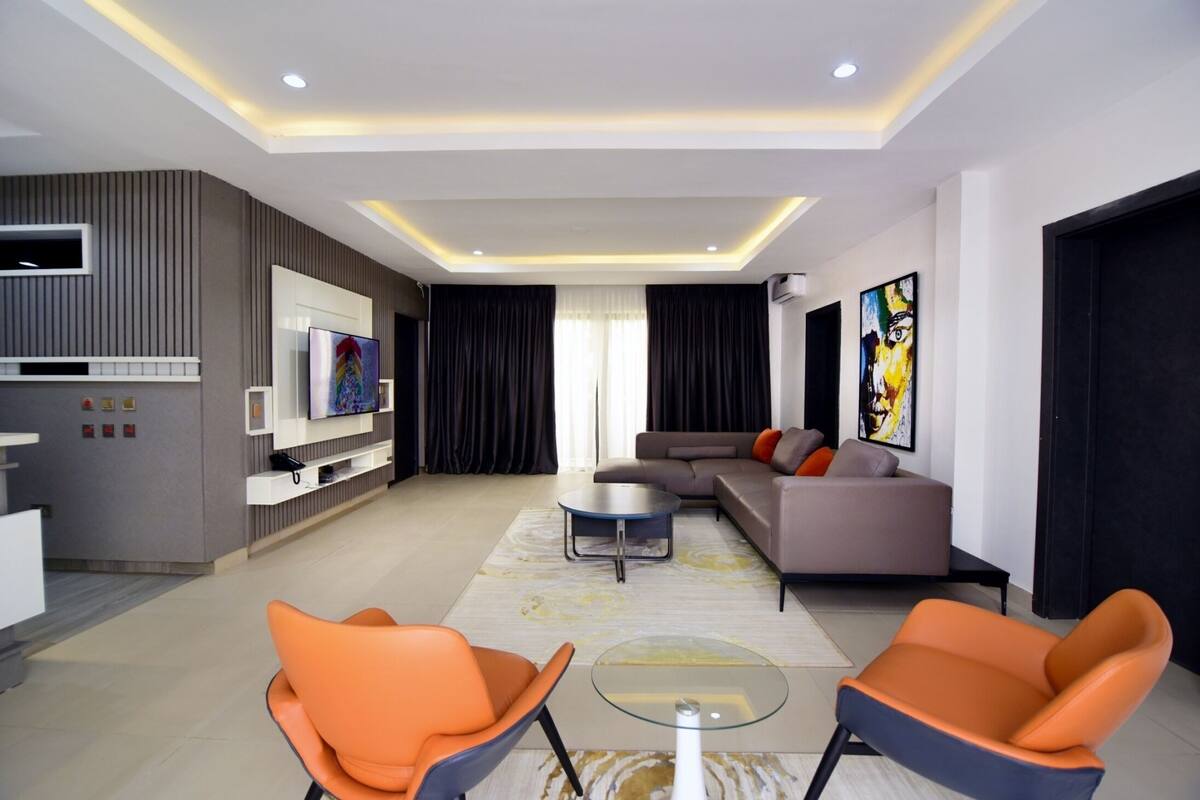 A modern living room with a sofa, two orange chairs, a glass coffee table, a flat-screen TV, and artwork on the walls.