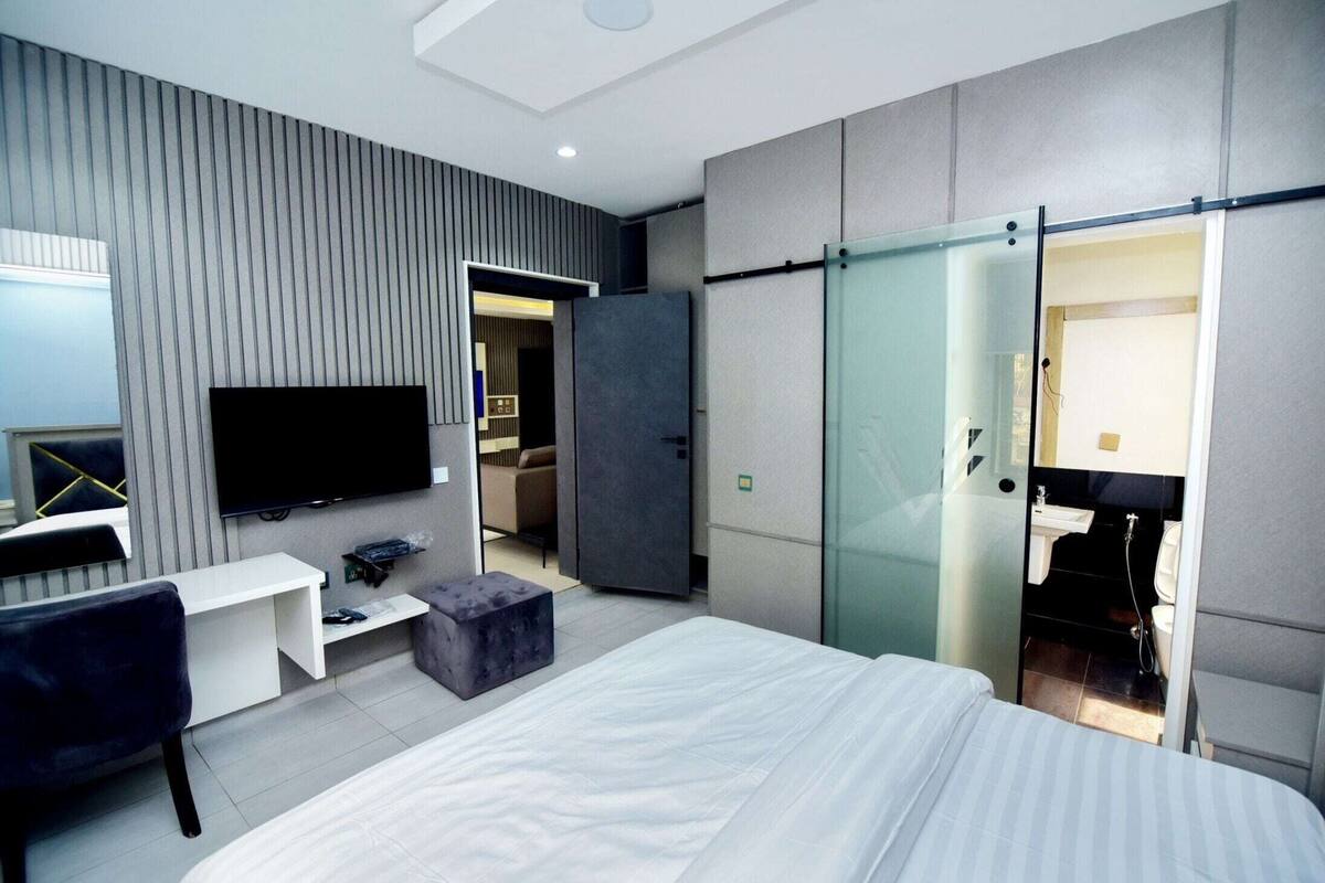 A modern hotel room with a bed, a desk, a flat-screen TV, a sliding door to a bathroom, and a view of the city.