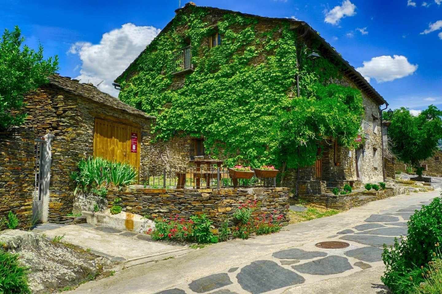 Ein Steinhaus mit einem Reetdach, umgeben von einem Kiesweg und üppiger Vegetation.