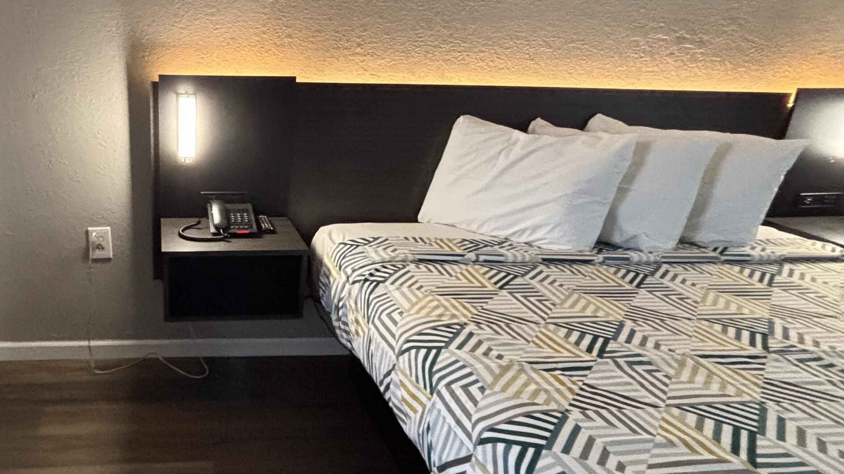 A hotel room with a bed, a bedside table with a phone, and a wall-mounted light.