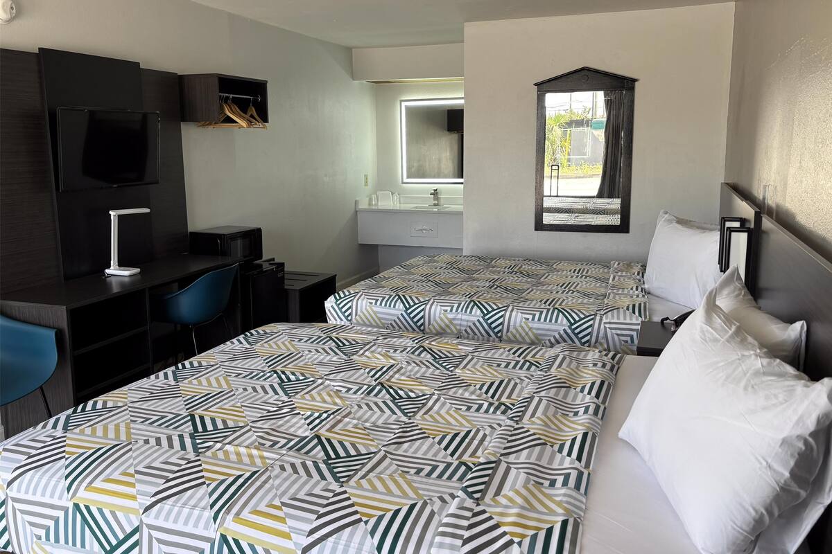 Une chambre d’hôtel avec deux lits, un bureau avec une chaise, une télévision, un miroir et une fenêtre donnant sur l’extérieur.