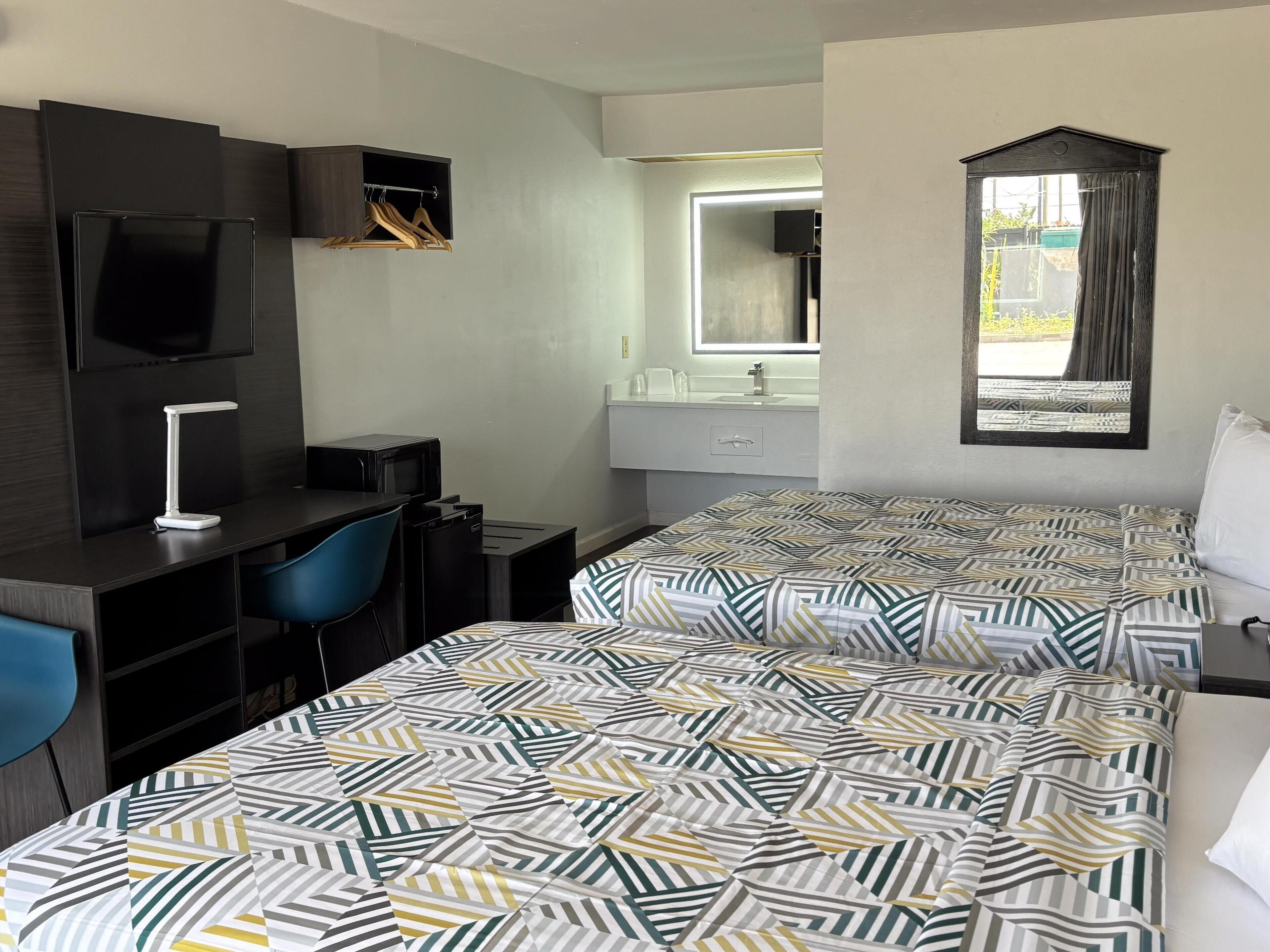 A hotel room with a bed, a desk with a chair, a TV, a mirror, and a window with curtains.