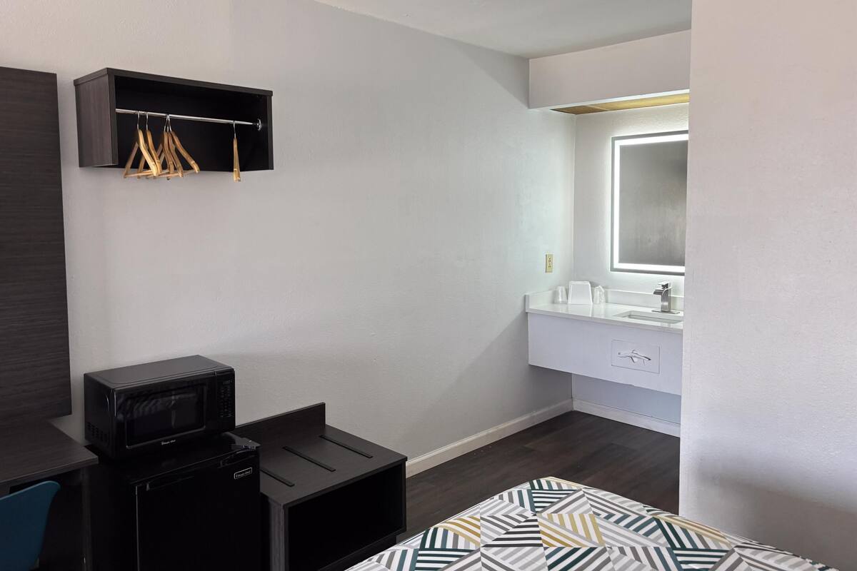 Une chambre d’hôtel moderne avec un plancher en bois foncé, un bureau noir doté d’un micro-ondes, une armoire avec une tringle, une coiffeuse blanche avec un miroir et une couvre-lit à motifs.