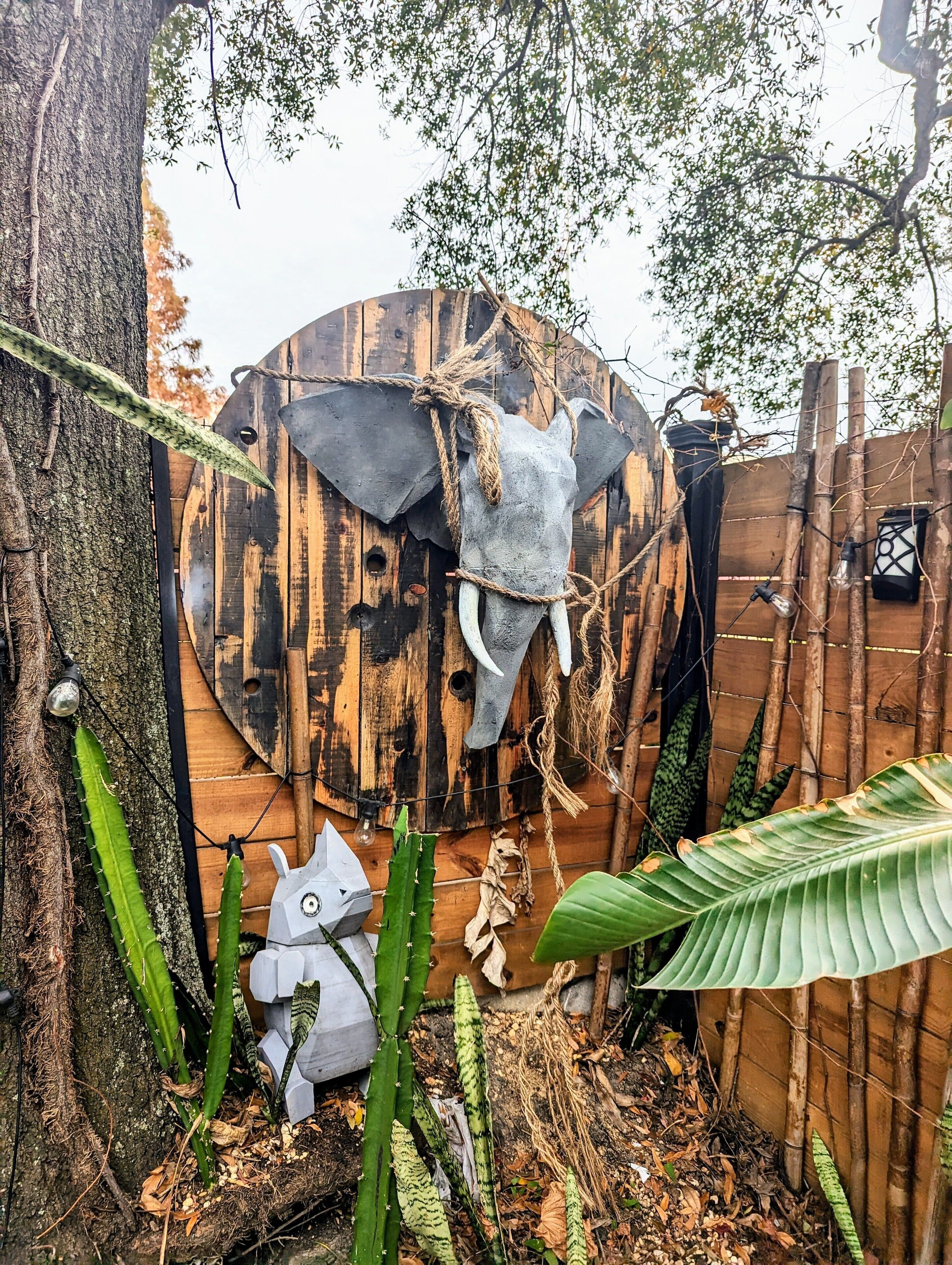 Une structure rustique en bois, surmontée d’une grande sculpture représentant une tête d’éléphant, entourée de verdure et délimitée par une clôture en bambou.