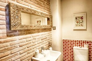 A bathroom with a brick wall, a white sink, a mirror with ornate frame, a toilet, and a framed floral artwork. - Le Chêne Creux Country House (Campillejo)