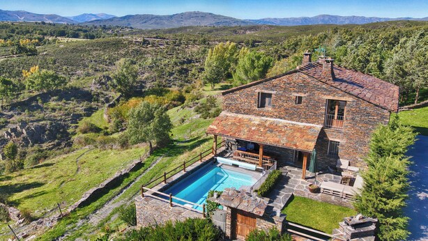 Une maison en pierre avec une piscine, entourée d’un paysage luxuriant et avec des montagnes en arrière-plan.