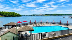 A swimming pool with lounge chairs and red umbrellas, surrounded by a fence, with a view of a lake and distant buildings. - The Lucky Minnow  • Pools, Hot Tub, Deck, Grill (Village of Four Seasons)
