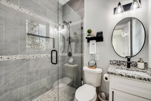 Une salle de bain moderne dotée d’une douche à parois de verre, de toilettes, d’un lavabo avec un plan de travail en marbre et d’un miroir rond.