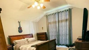 Un dormitorio con cama, ventilador de techo, guitarra en la pared, una cruz y televisor.